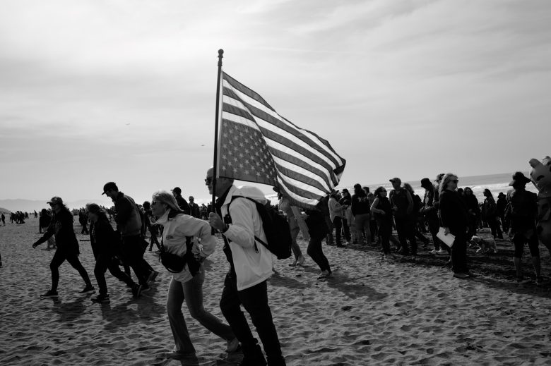 A group of people walk on a sandy beach; one person in the foreground carries a large American flag.