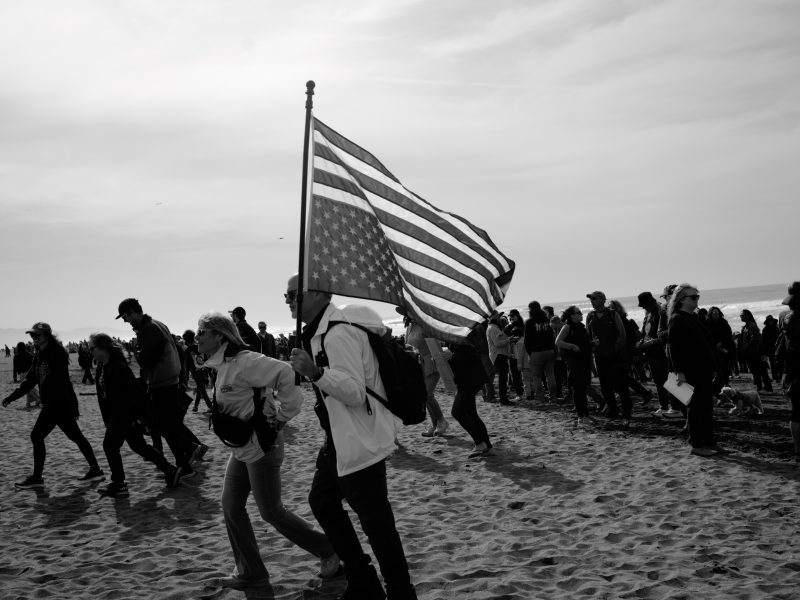 A group of people walk on a sandy beach; one person in the foreground carries a large American flag.