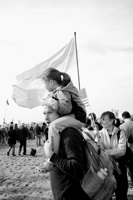 A man carries a child on his shoulders at a crowded outdoor event, with several people holding flags in the background.