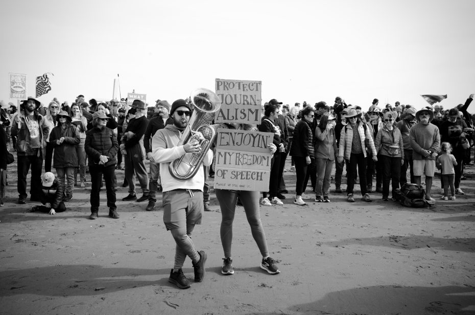 A person holds a sign reading "Protect Journalism, Enjoying My Freedom of Speech" while another plays a tuba; a crowd stands behind them on a beach.