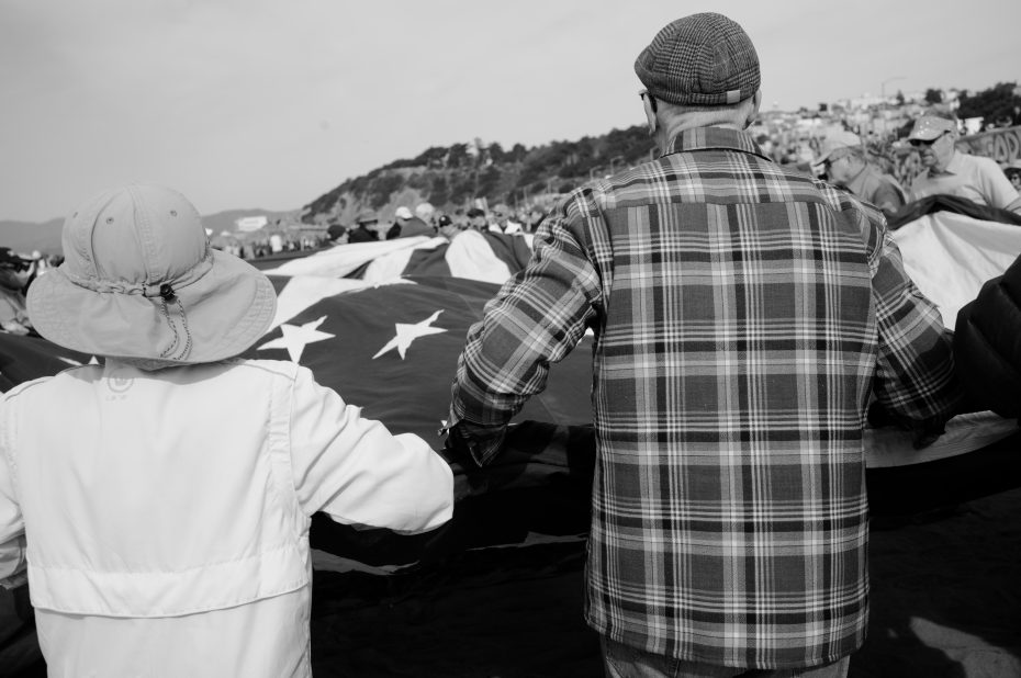 Two people, seen from behind, hold a large American flag along with a group outdoors. The photo is in black and white.