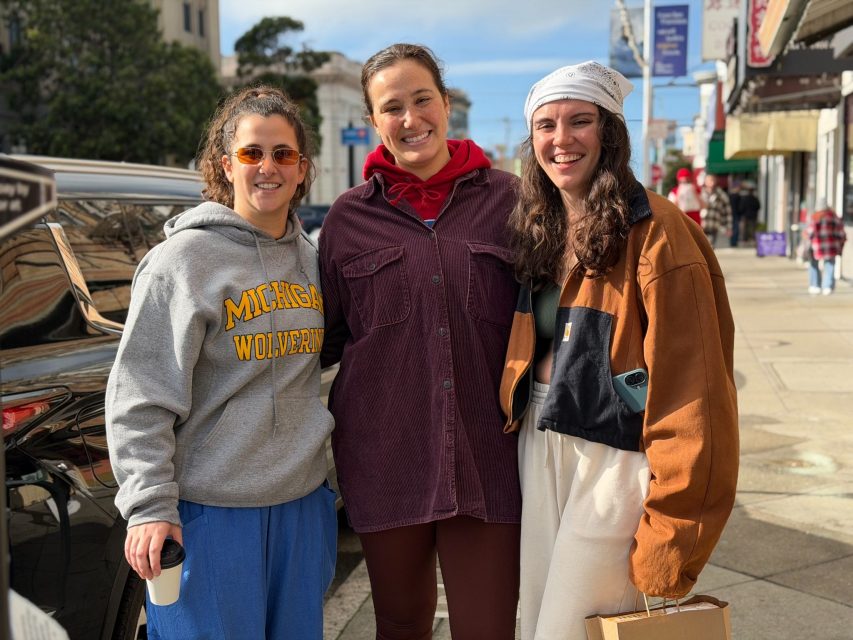 Three women standing and smiling on a city sidewalk; one wears a Michigan Wolverines sweatshirt, another a red hoodie, and the third a brown jacket and white headscarf.