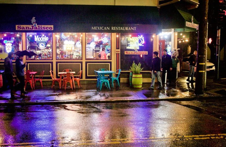 Exterior view of a Mexican restaurant at night with neon signs, colorful outdoor chairs and tables, and people walking on a wet sidewalk.
