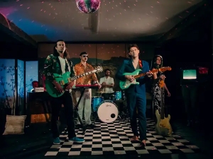 A five-member band performs on a black-and-white checkered floor under a disco ball, with drums, electric guitars, and a keyboard visible.
