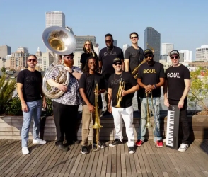 A group of ten musicians with brass instruments and a keyboard pose together on a rooftop with a city skyline in the background.
