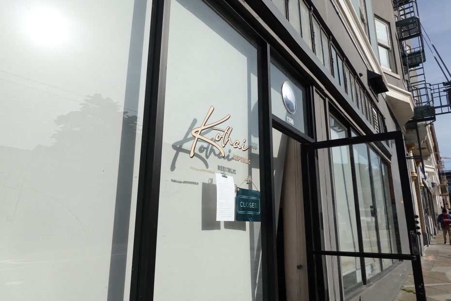 Street view of a storefront with "Kohari" signage, a "CLOSED" sign, and a notice posted on the glass door; the building exterior is modern and minimalist.