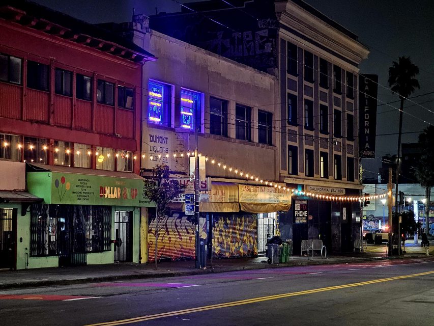 A city street at night with closed shops, neon signs in windows, string lights, and colorful graffiti on building fronts.