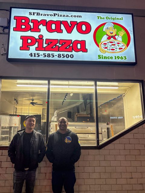 Two men stand outside Bravo Pizza at night under a brightly lit sign displaying the restaurant's name, phone number, and "The Original Since 1963.