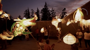 People hold illuminated lanterns shaped like animals outdoors at dusk, with trees and a wooden building in the background.