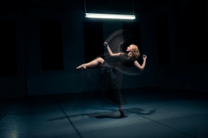 A woman in a black dress performs a dance move under a bright overhead light in a dimly lit room, with motion blur showing movement.
