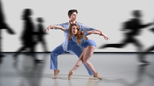 Two ballet dancers in blue outfits pose in a dance studio while blurred figures rush past them, highlighting a contrast between stillness and motion.
