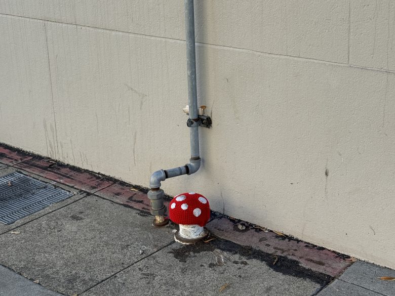 A metal pipe runs along a beige wall, with a fire hydrant cap covered in red yarn with white spots, resembling a mushroom, on a concrete sidewalk.