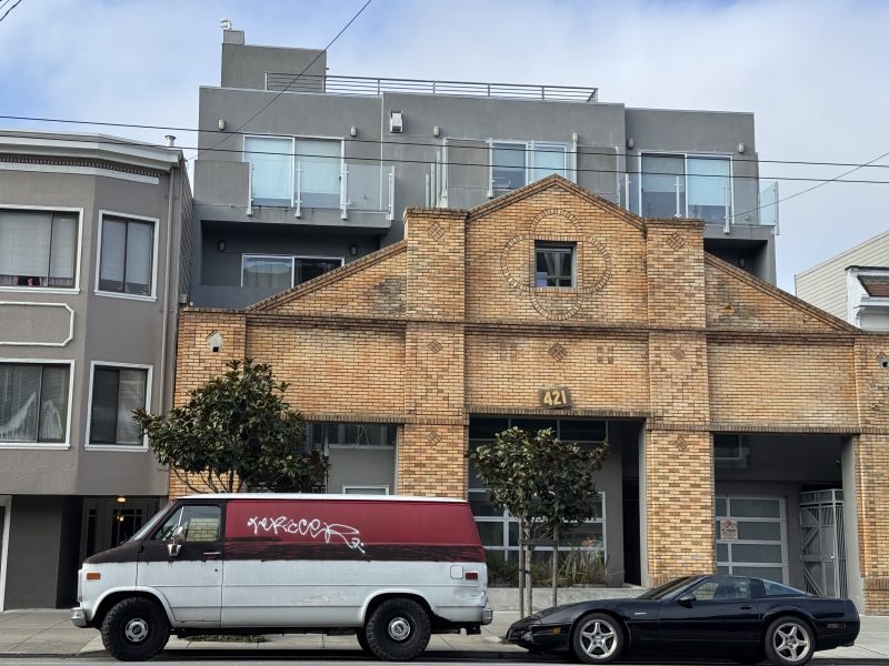 A brick building with modern glass balconies stands behind a red-and-white van with graffiti and a black sports car parked on the street.