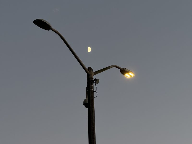 A streetlamp with one light on and one off stands against a dusky sky, with a half-moon visible in the background.