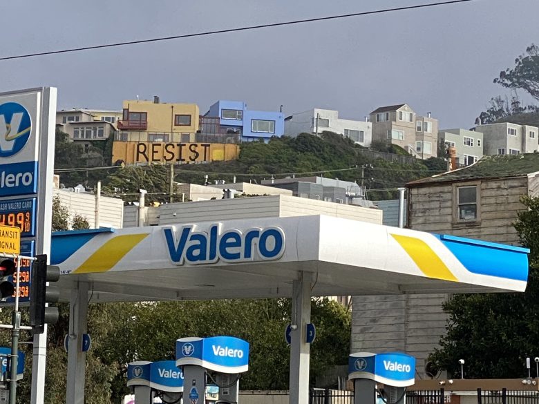 A Valero gas station in the foreground with houses on a hillside behind, one of which has large letters spelling "RESIST" on its facade.
