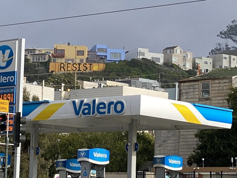 A Valero gas station in the foreground with houses on a hillside behind, one of which has large letters spelling "RESIST" on its facade.