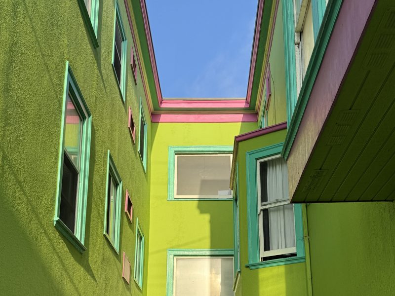 Two brightly painted buildings in yellow, green, and pink are separated by a narrow alley, with windows and a blue sky visible above.