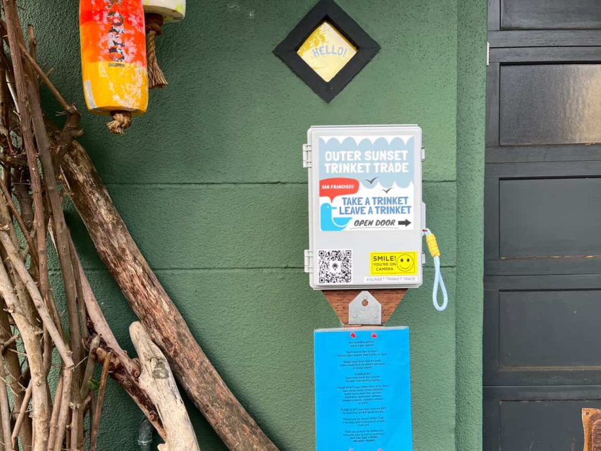 A small outdoor box labeled "Outer Sunset Trinket Trade" invites people to take or leave a trinket; a blue notice and various decorations are mounted on a green wall nearby.