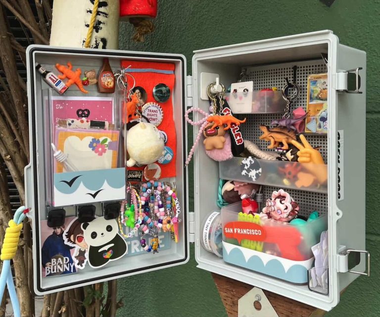 Two open wall-mounted boxes display an assortment of small toys, keychains, stickers, mini-figures, pins, and colorful trinkets against a green background.