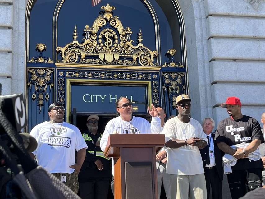 A group of men stand in front of a podium outside City Hall; one man speaks while others stand nearby wearing "United in Peace" T-shirts.