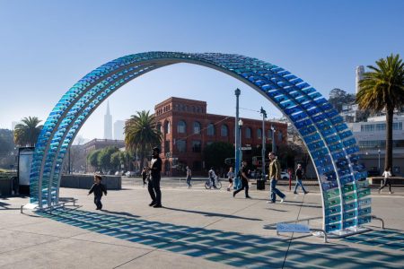 People walk and play near a blue arched art installation in a sunny urban plaza, surrounded by palm trees and red brick buildings, not far from nearby museums.
