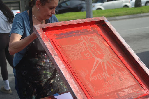 A person participates in an ICE walkout, using a screen printing frame with red ink outdoors to create a design on fabric, while cars are parked on the street in the background.