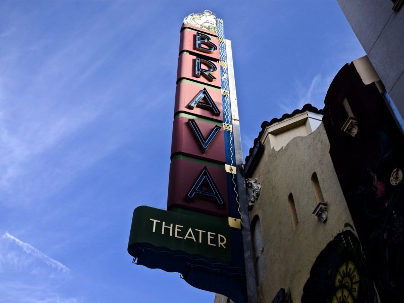 Vertical sign reading "BRAVA THEATER" on the exterior of a building, photographed from below against a blue sky.