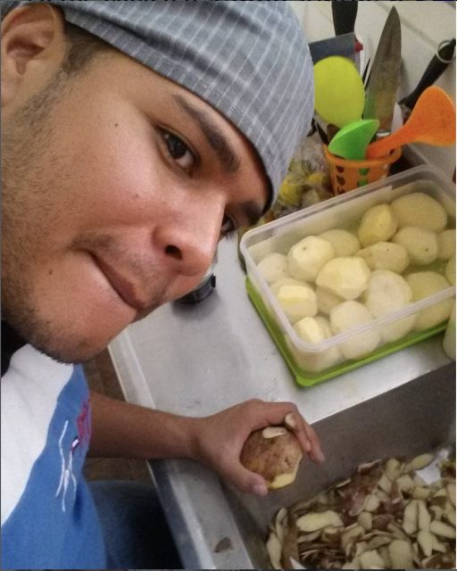 A person wearing a gray cap is peeling potatoes at a kitchen counter, with peeled potatoes and kitchen utensils visible in the background.