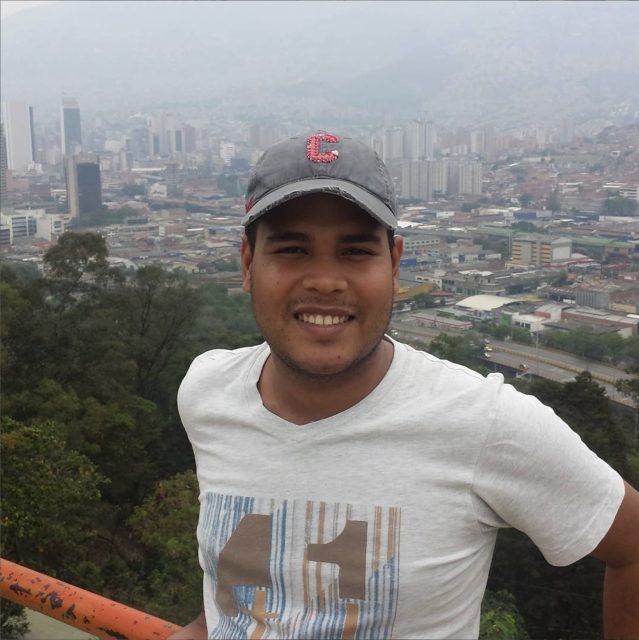 A man wearing a gray cap and white t-shirt stands by a railing with a cityscape and greenery in the background on a cloudy day.
