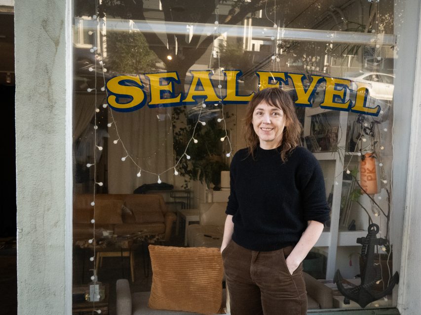 A person with brown hair stands in front of a window displaying the word "SEALEVEL"; indoor seating and plants are visible inside.