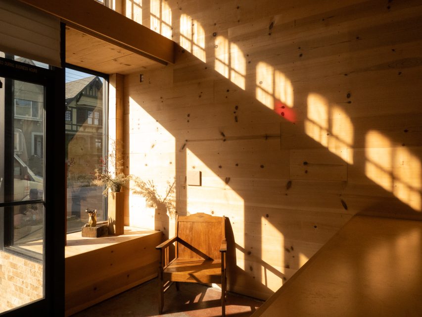 Sunlight streams through windows, casting shadows on a wooden wall in a cozy room with a chair, small table, and dried flowers by the window.