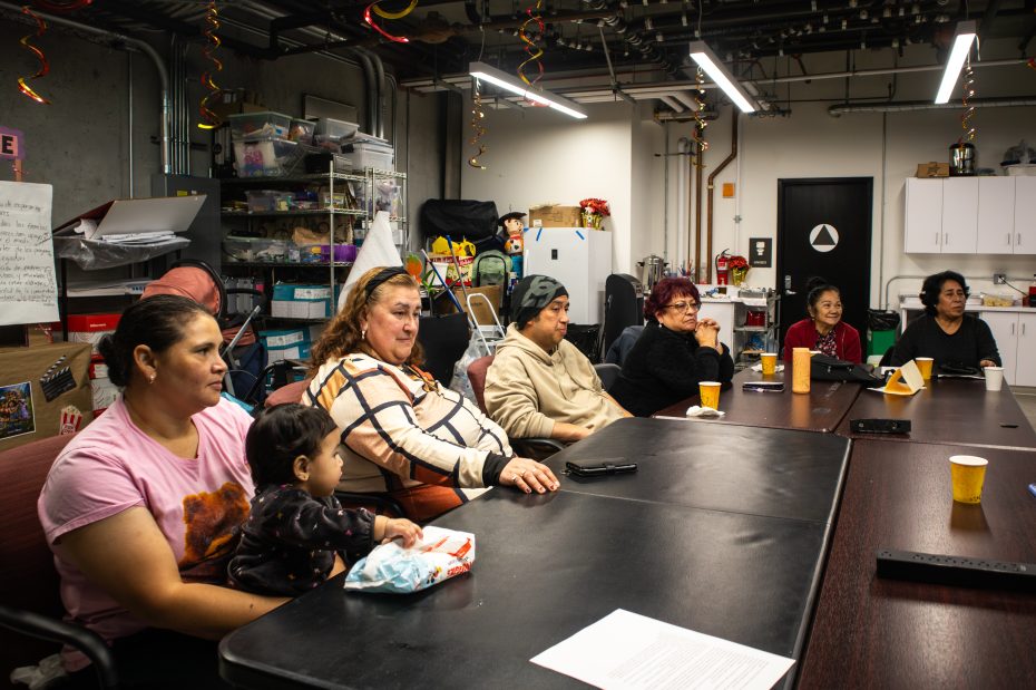 Seven adults and one child sit around a conference table in a cluttered room, appearing to listen attentively during a meeting.