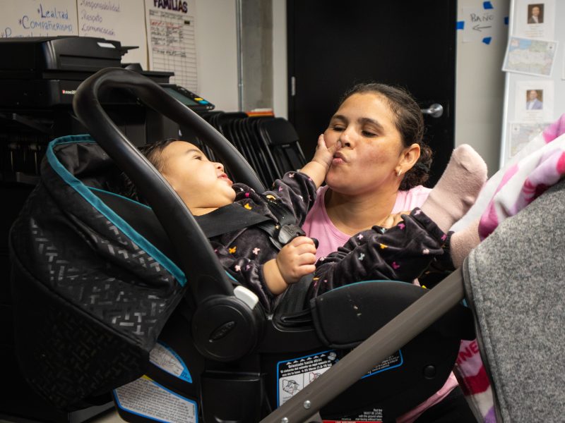 A woman leans toward a baby in a car seat as the baby touches her face, both smiling; office supplies and papers are visible in the background.