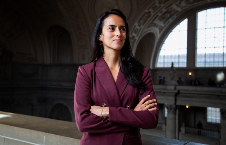 A woman in a maroon blazer stands with arms crossed indoors, in front of large arched windows and ornate architectural details.