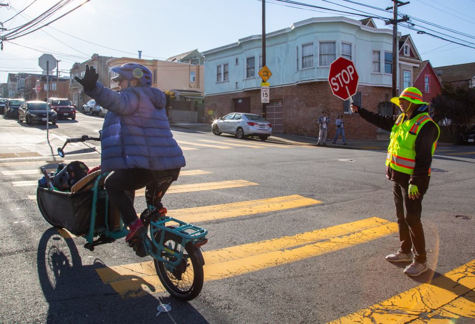 A crossing guard holds a stop sign while a cyclist with a child in a rear seat raises a hand, crossing a sunny street intersection.