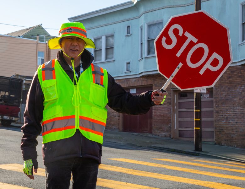 A crossing guard in a neon safety vest and hat holds a stop sign at a crosswalk on a residential street.