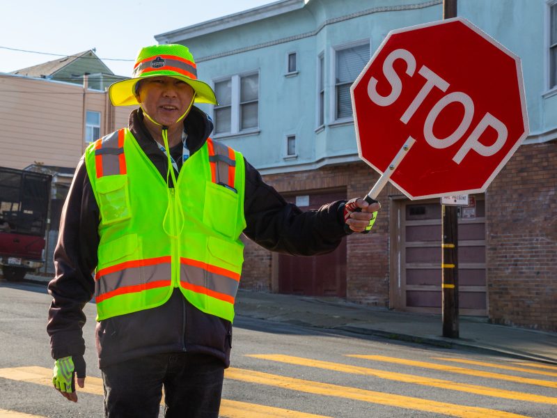 A crossing guard in a neon safety vest and hat holds a stop sign at a crosswalk on a residential street.