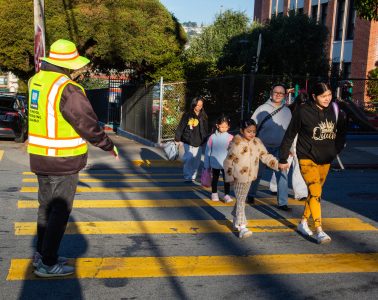 A crossing guard in a reflective vest helps a group of children and adults cross a yellow-striped crosswalk on a sunny day.