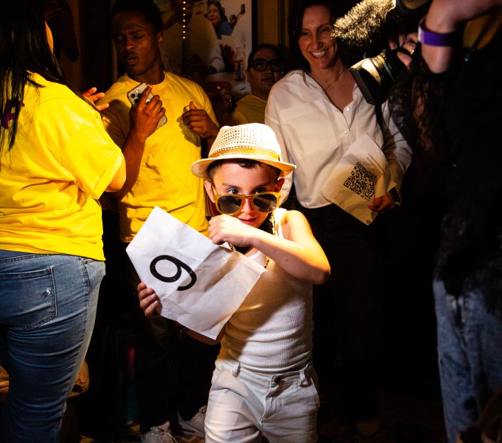 A young child in sunglasses and a hat holds a paper with the number 9, surrounded by people, some wearing yellow shirts, at an indoor event.