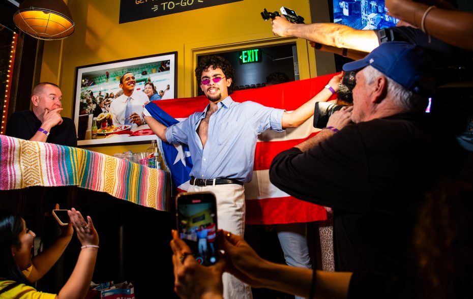 A person poses holding a Puerto Rican flag in front of cameras and phones in a brightly lit room with photos on the wall behind them.