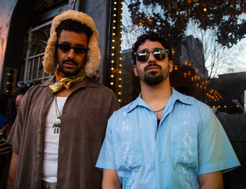 Two men wearing sunglasses and casual shirts stand outside a building with string lights, one in a brown shirt with a fur hat, the other in a blue shirt with embroidery.