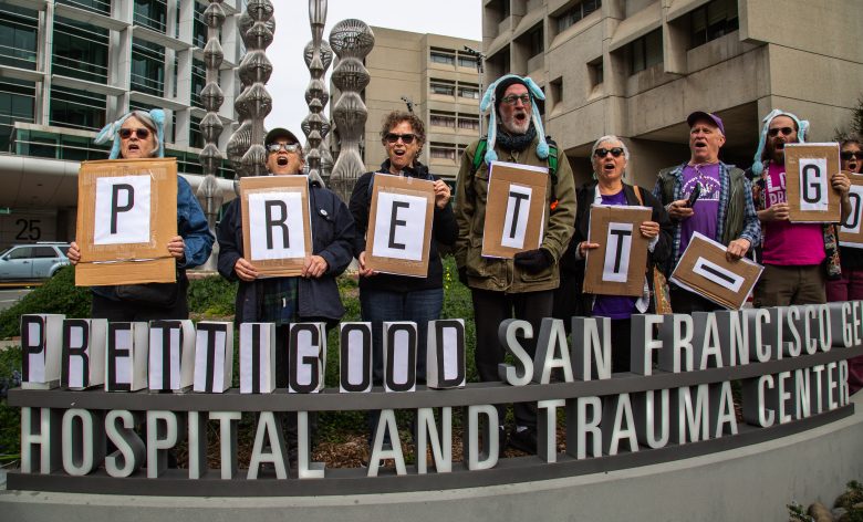 A group of people stand outside a hospital holding large signs that collectively spell "PRETTY GOOD" above the San Francisco General Hospital and Trauma Center sign.