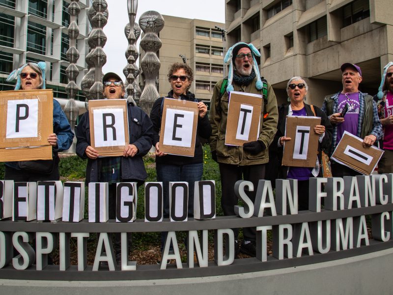 A group of people stand outside a hospital holding large signs that collectively spell "PRETTY GOOD" above the San Francisco General Hospital and Trauma Center sign.