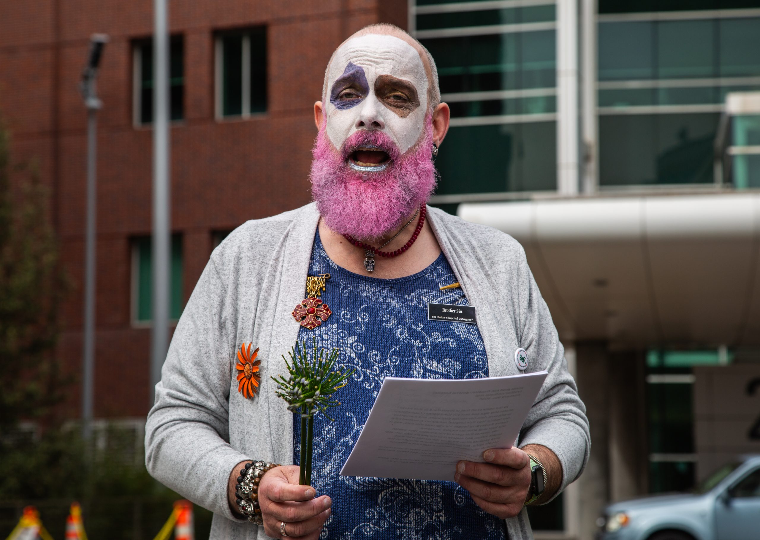 Person with a painted face and pink beard holds papers and a small plant, standing outdoors in front of a modern building.