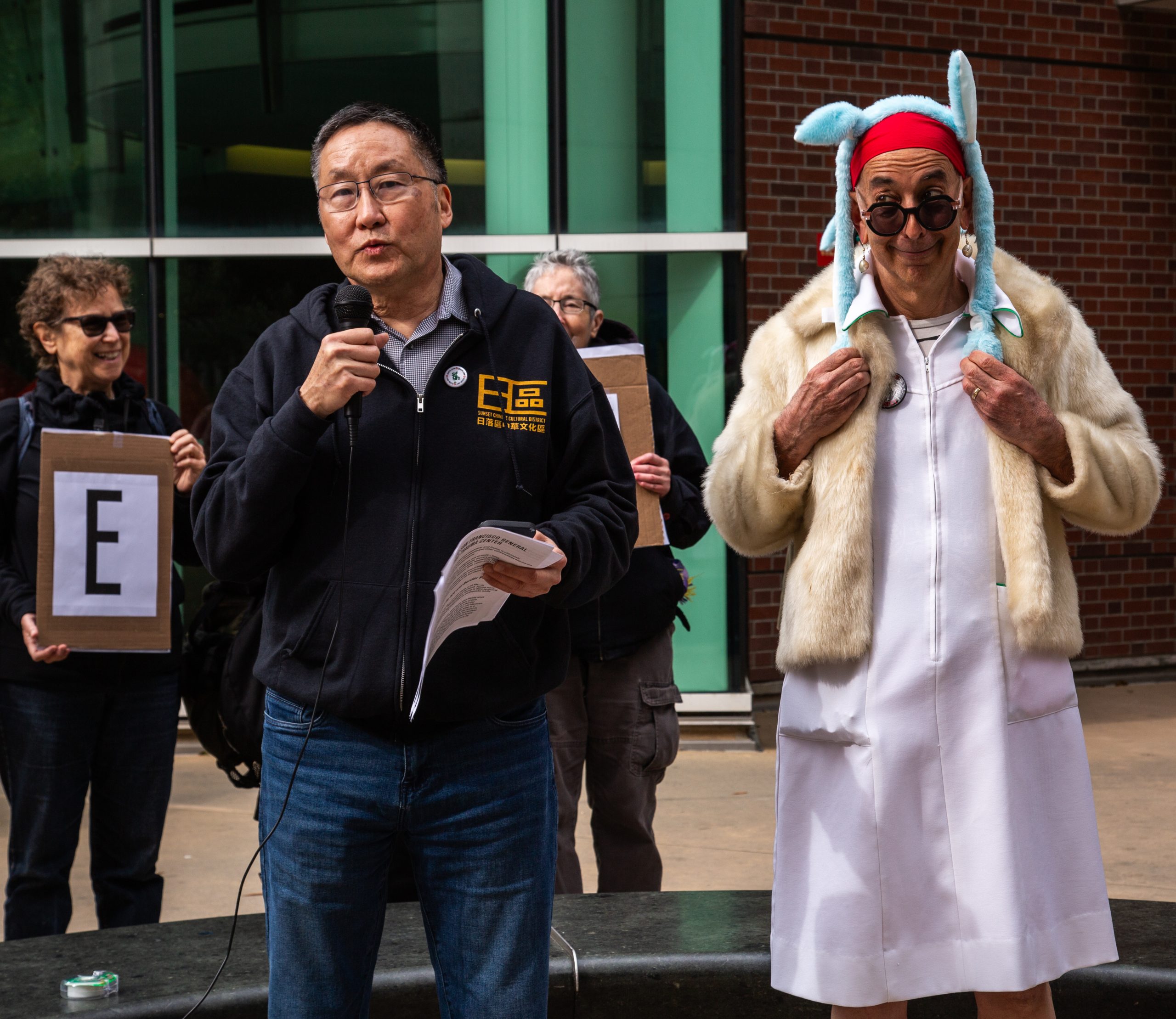 A man speaks into a microphone while another person in a white coat and blue hat stands beside him; two others hold letter "E" signs in the background.