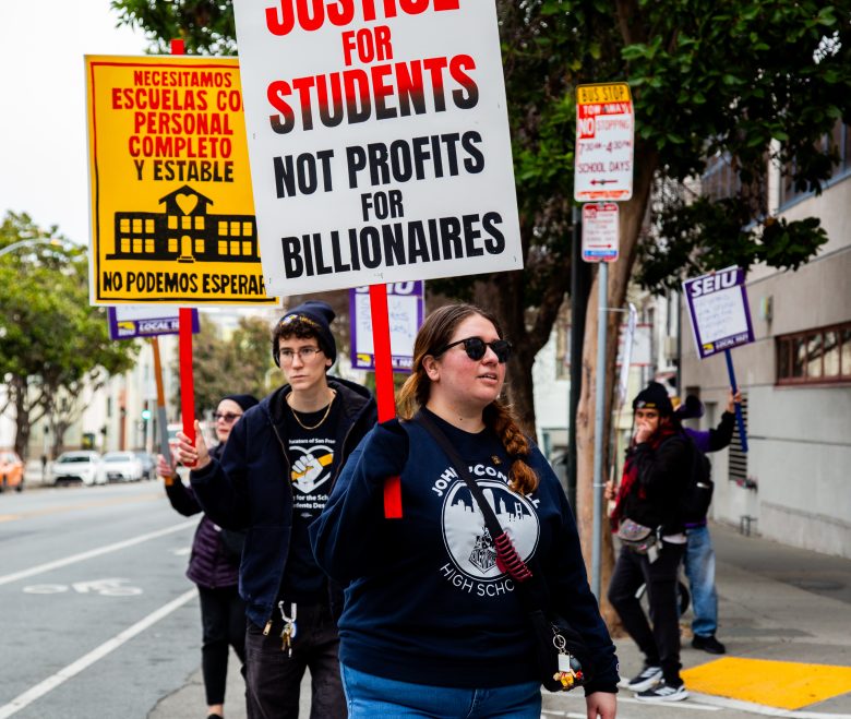 A group of people march on a city sidewalk holding protest signs advocating for justice for students and more school staff, with one sign in Spanish.