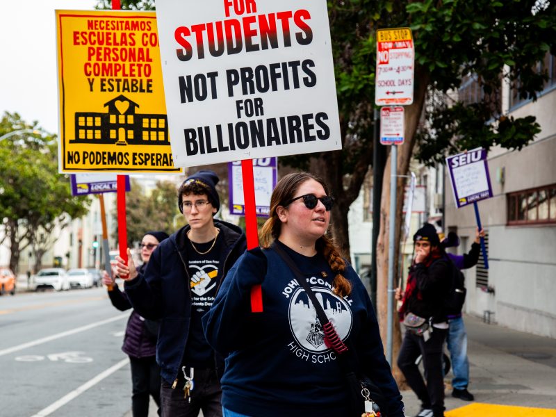 A group of people march on a city sidewalk holding protest signs advocating for justice for students and more school staff, with one sign in Spanish.