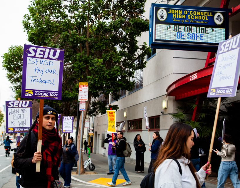 Protesters holding SEIU signs supporting teachers walk past John O'Connell High School, with a marquee that reads "Be On Time! Be Safe.