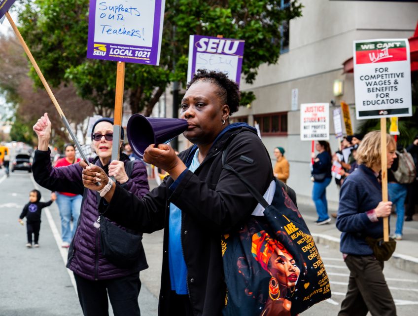 A group of people march on a city street holding protest signs in support of teachers and better wages for educators. One person speaks into a megaphone.