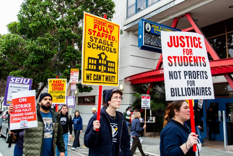 A group of people participate in a protest, carrying signs demanding well-staffed schools, student justice, and fully funded healthcare.